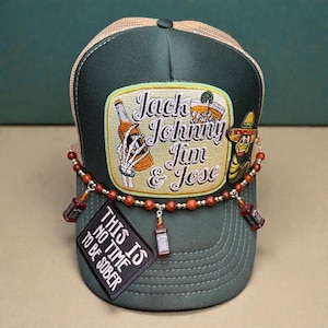 May include: A green and tan trucker hat with the embroidered text "Jack Johnny Jim & Jose". The hat features a patch with a skeleton holding a bottle and a patch with a worm wearing a sombrero. A beaded chain with miniature bottle charms hangs from the brim. The text "THIS IS NO TIME TO BE SOBER" is on a patch.