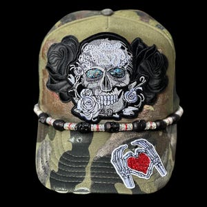 May include: A camouflage baseball cap featuring a skull design with black roses and a heart-shaped hand gesture. The skull has glittery blue eyes and is surrounded by white roses. A beaded band accents the brim.