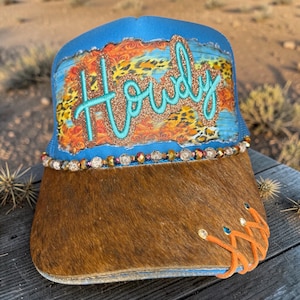 May include: A blue trucker hat with the word "Howdy" in turquoise script. The hat features a brown fur brim, a decorative beaded band, and a colorful design with leopard print and orange accents.