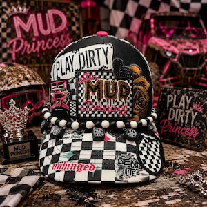 May include: A black and white checkered baseball cap featuring patches that read PLAY DIRTY and MUD PRINCESS. The cap is adorned with a string of black and white beads with miniature tire charms, a black heart patch, and various graphic prints.