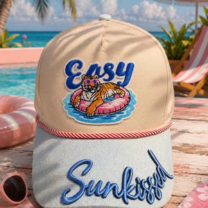 May include: A beige and light blue baseball cap featuring a red and white twisted cord trim. The front panel displays an embroidered patch of a tiger wearing pink sunglasses on a pink pool float with the word Easy. The brim features Sunkissed.