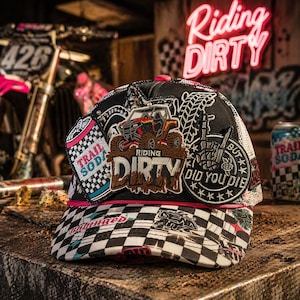 May include: Black trucker hat with a checkered brim and patches. The patches include the words "Riding Dirty" and "Trail Soda", along with a graphic of an off-road vehicle. The hat has a mesh back.