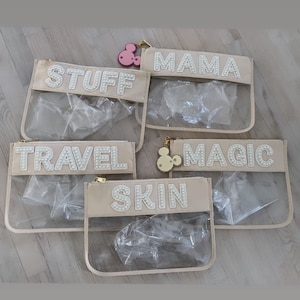 May include: Five clear plastic zippered pouches with beige fabric trim. Each pouch has a different word spelled out in white pearl-like beads: "Stuff", "Mama", "Travel", "Magic", and "Skin".