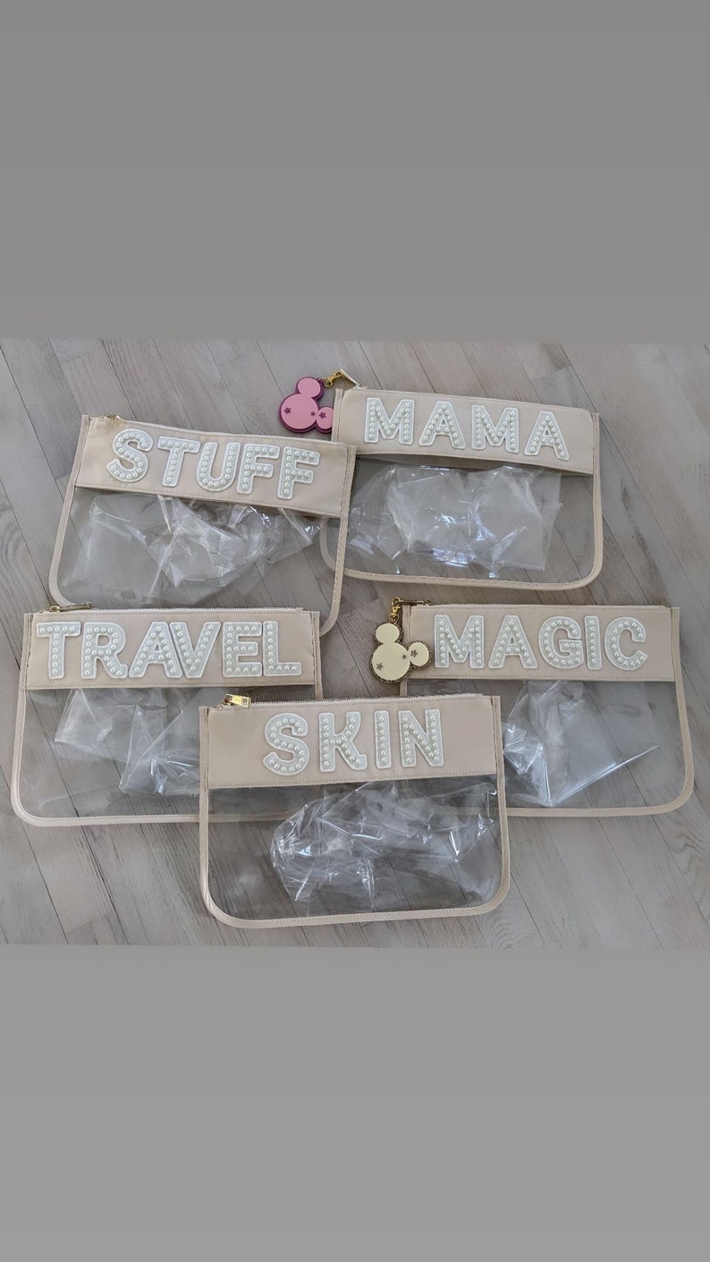 May include: Five clear plastic zippered pouches with beige fabric trim. Each pouch has a different word spelled out in white pearl-like beads: "Stuff", "Mama", "Travel", "Magic", and "Skin".