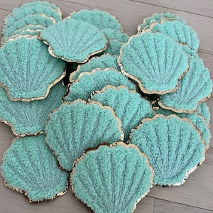 May include: A collection of aqua-colored seashell-shaped coasters with gold trim. The coasters have a textured, plush appearance and are arranged on a light-colored wooden surface. The design evokes a coastal or beach theme.