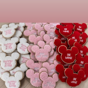 May include: A collection of handmade felt Mickey Mouse-shaped ornaments. The ornaments are in three colors: white, pink, and red. The white ornaments have the text "Happily Ever After" embroidered on them. The pink ornaments have the text "Best Day Ever" embroidered on them. The red ornaments have the text "Be Mine" embroidered on them.