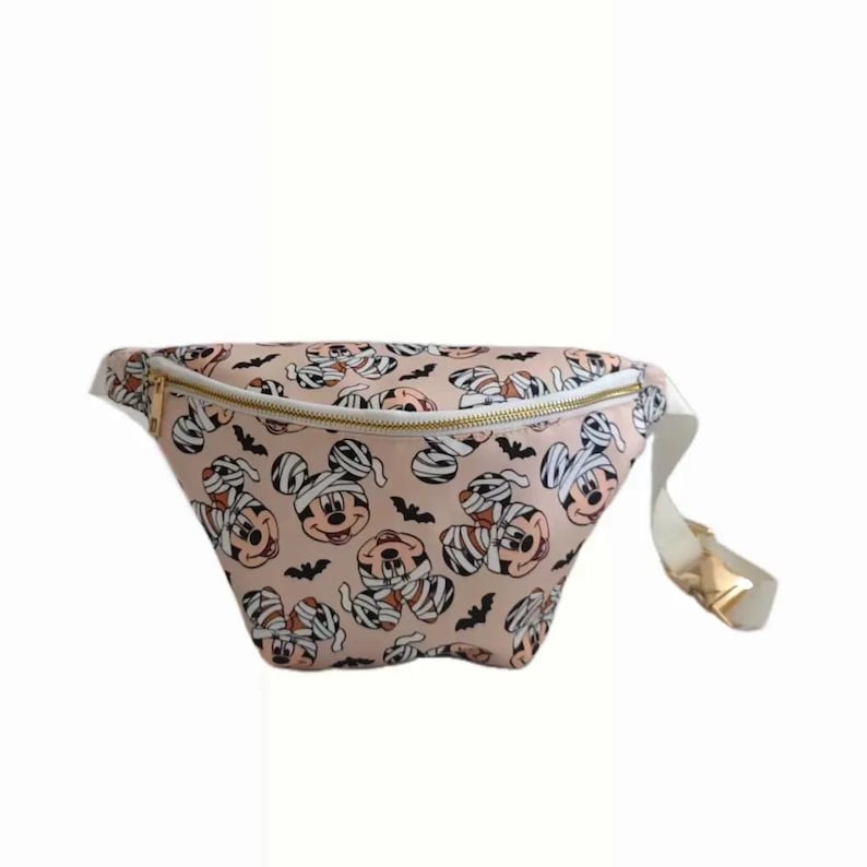 May include: A pink fanny pack with a white adjustable strap. The fanny pack has a gold zipper and a print of Mickey Mouse dressed as a mummy with bats.