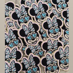 May include: A pile of black, pink, and gold embroidered iron-on patches featuring a cartoon character with a bow, sunglasses, and a star pattern.
