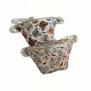 May include: Two Halloween themed fanny packs with a Mickey Mouse mummy design. The fanny packs are white and pink with black bats and gold zippers.