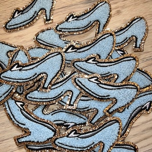 May include: A collection of light blue high-heeled shoe patches with gold glitter trim and black accents. The patches are scattered on a light wood surface. The shoes have a textured appearance.