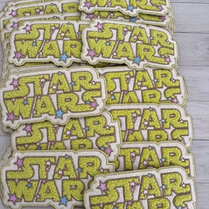 May include: A stack of yellow and white embroidered patches with the text "STAR WARS" in a retro font. The patches are decorated with pink and blue stars.