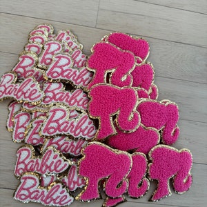 May include: A collection of pink and gold embroidered patches. The patches include the word "Barbie" in a cursive font and the silhouette of a woman's head with a ponytail. The patches have a gold border.