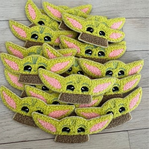 May include: A pile of green, brown, and pink felt patches shaped like a popular fictional character. The patches are all the same design, with large eyes, small mouths, and large ears.