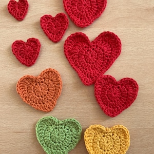 May include: A set of seven crocheted hearts in various sizes and colours, including red, orange, yellow, and green. The hearts are arranged on a wooden surface.