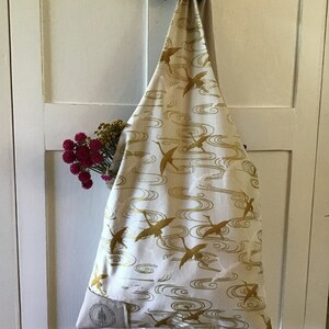 May include: A white fabric tote bag with a gold crane and wave pattern. The bag is hanging on a white door.