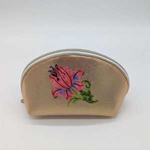 May include: A gold-coloured, half-circle shaped cosmetic bag with a white zipper and a pink and green embroidered flower design.