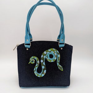 May include: A blue denim handbag with a light blue handle and a green and blue snake design embroidered on the front.