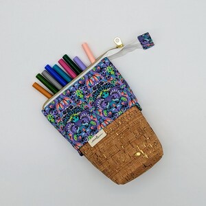 May include: A cork and fabric pencil case with a zipper closure. The fabric is purple with a colorful floral pattern. The cork is brown with gold flecks. The case is filled with colored markers.