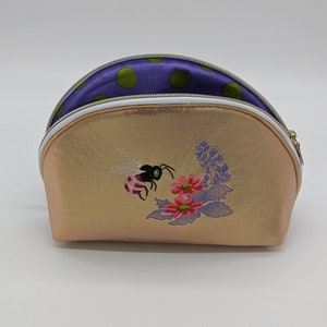 May include: A gold-colored cosmetic bag with a white zipper and a bee and flower embroidery design. The inside lining is purple with green polka dots.