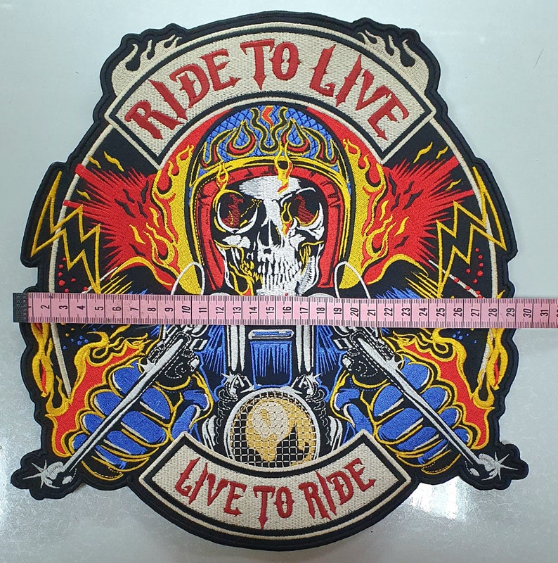May include: Embroidered patch featuring a skull wearing a helmet, flames, and the text "RIDE TO LIVE" and "LIVE TO RIDE". The patch is approximately 12 inches wide. The design includes a motorcycle and crossed weapons, with a color palette of red, yellow, blue, and black.