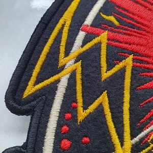 May include: Embroidered patch with a black background and a design featuring yellow lightning bolts, red bursts, and white accents. The patch has a black border and is likely for clothing or accessories.
