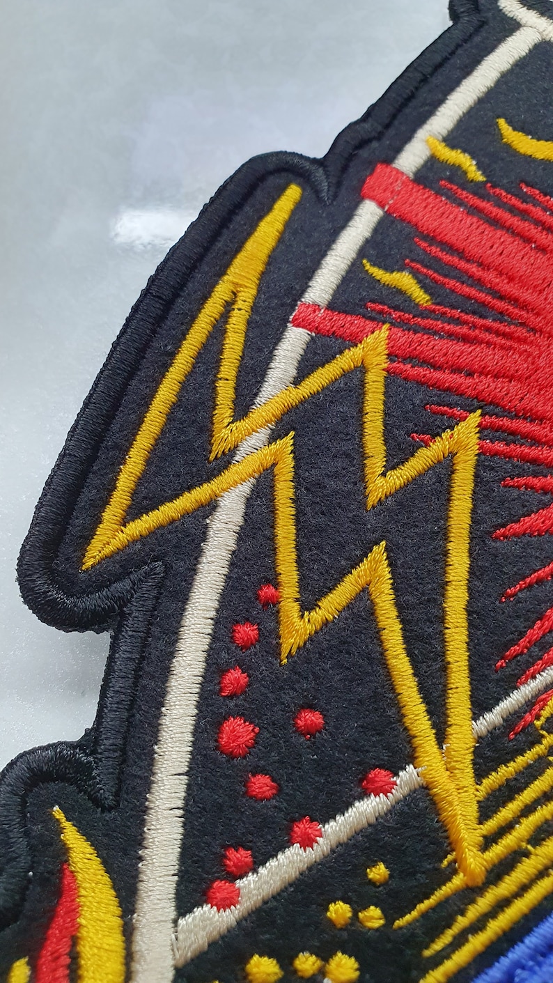 May include: Embroidered patch with a black background and a design featuring yellow lightning bolts, red bursts, and white accents. The patch has a black border and is likely for clothing or accessories.