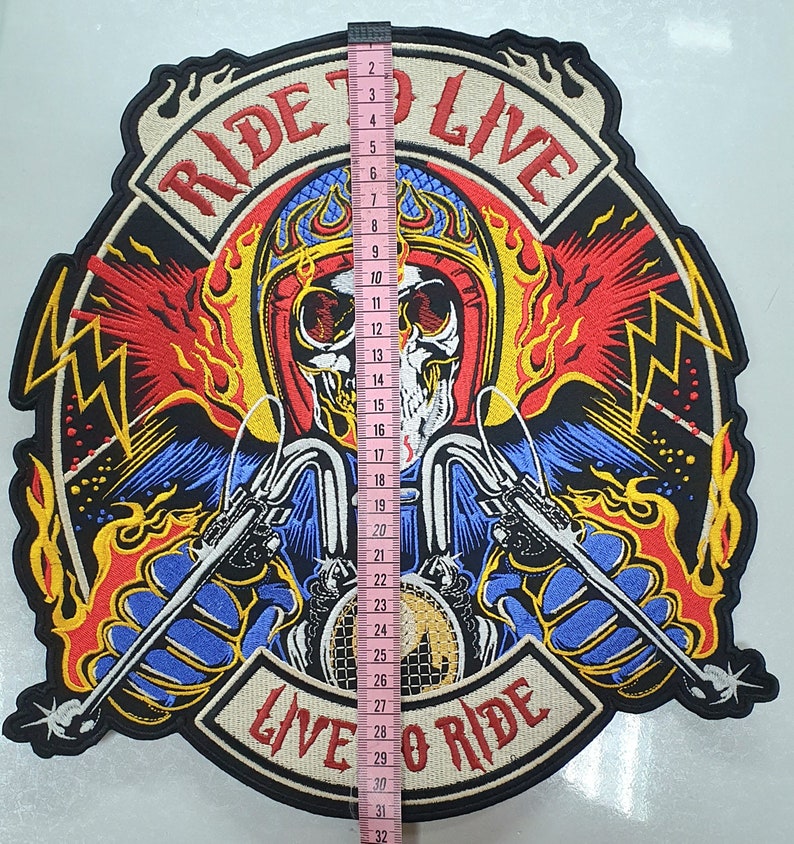 May include: Embroidered patch featuring a skull riding a motorcycle, with the text "RIDE TO LIVE" and "LIVE TO RIDE". The patch is approximately 12 inches tall. The design incorporates red, yellow, blue, and black colors, with a detailed, flame-like background.