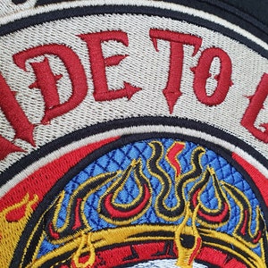 May include: Embroidered patch with the words "RIDE TO LIVE" in red gothic lettering. The patch features a flame design in yellow, red, and orange, with a blue and gold patterned center. The patch is bordered in black and beige.