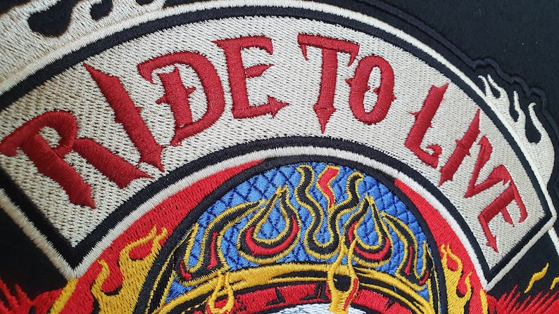 May include: Embroidered patch with the words "RIDE TO LIVE" in red gothic lettering. The patch features a flame design in yellow, red, and orange, with a blue and gold patterned center. The patch is bordered in black and beige.