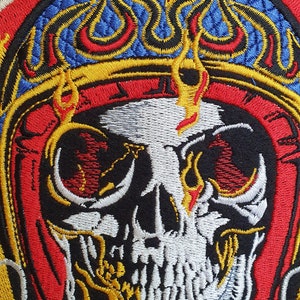 May include: Embroidered patch featuring a detailed skull design with flames and intricate stitching. The skull is white with black accents, surrounded by red, blue, and yellow flames and borders. The patch has a textured background.