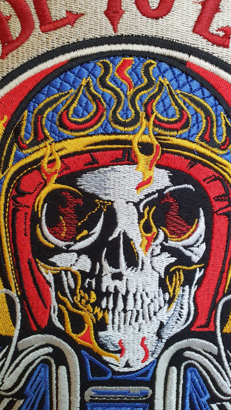 May include: Embroidered patch featuring a detailed skull design with flames and intricate stitching. The skull is white with black accents, surrounded by red, blue, and yellow flames and borders. The patch has a textured background.