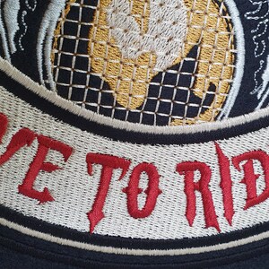 May include: Embroidered patch with the words "LIVE TO RIDE" in red, gothic-style lettering. The patch features a black border, cream-colored background, and a detailed design with gold, blue, and white accents. The patch is likely for a motorcycle jacket or vest.