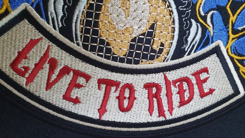May include: Embroidered patch with the words "LIVE TO RIDE" in red, gothic-style lettering. The patch features a black border, cream-colored background, and a detailed design with gold, blue, and white accents. The patch is likely for a motorcycle jacket or vest.