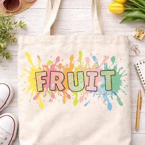 以下が含まれることがあります： クリーム色のトートバッグに、カラフルな大きな文字で「FRUIT」の文字が描かれています。様々な色のペイントスプラッターを背景に、バッグには2つのハンドルがあり、麦わら帽子、サングラス、花に囲まれています。
