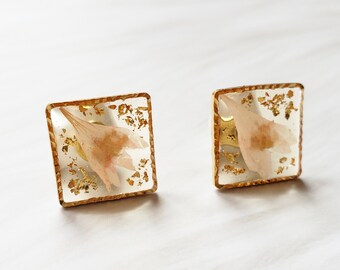 Könnte beinhalten: Ein Paar goldene quadratische Ohrringe mit einem zarten Design aus gepressten Blumen. Die Blumen sind hellrosa und in klarem Harz gefasst.