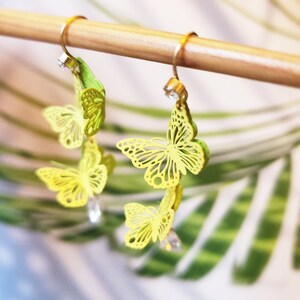 Pode incluir: Um par de brincos de borboleta dourados com detalhes em amarelo e verde. Os brincos estão pendurados em uma haste de madeira.