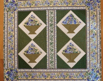 May include: A green and white quilt with a floral pattern. The quilt features four diamond-shaped panels with a basket of flowers in each panel. The quilt is bordered with a floral print.