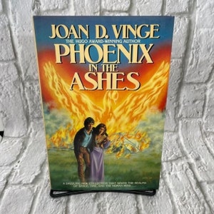 May include: A book cover with a fiery phoenix design. The title is "Phoenix in the Ashes" by Joan D. Vinge, a Hugo Award-winning author. The text on the cover reads "A dazzling new collection that spans the realms of space, time, and the human mind..."