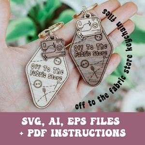 Op de afbeelding: Twee houten sleutelhangers in de vorm van vintage motelborden met de tekst "Off To The Fabric Store" en een naaimachinegrafiek. De sleutelhangers worden in een hand gehouden.