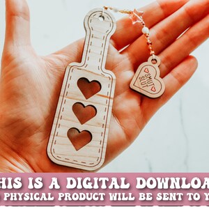 May include: Wooden paddle with three heart cutouts and a small heart-shaped charm that says "SMUT SLUT". The paddle is light brown with a stitched border. The charm is attached to a beaded strand. Text at the bottom reads "THIS IS A DIGITAL DOWNLOAD NO PHYSICAL PRODUCT WILL BE SENT TO YOU".