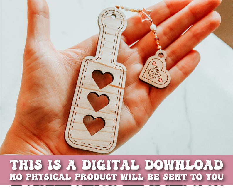 May include: Wooden paddle with three heart cutouts and a small heart-shaped charm that says "SMUT SLUT". The paddle is light brown with a stitched border. The charm is attached to a beaded strand. Text at the bottom reads "THIS IS A DIGITAL DOWNLOAD NO PHYSICAL PRODUCT WILL BE SENT TO YOU".