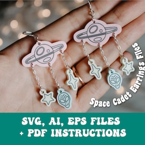 May include: A pair of pink and blue space cadet earrings featuring a planet, stars, and alien faces. The earrings are displayed on a hand, with the text "Space Cadet Earrings Files" visible in the background.