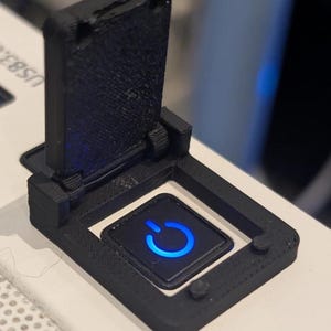 May include: A black 3D-printed power button cover with a hinged lid. The button has a blue illuminated power symbol. The cover is mounted on a white surface.