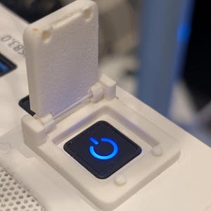 May include: A white, 3D-printed power button cover with a hinged lid. The button is black with a blue power symbol illuminated. The cover is open, revealing the button. The background is a white surface.