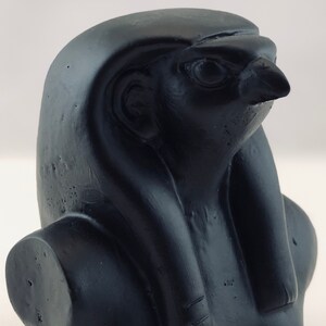 May include: Black resin bust of the Egyptian god Horus, with a falcon head and a human body. The bust is detailed with a headdress and a collar.