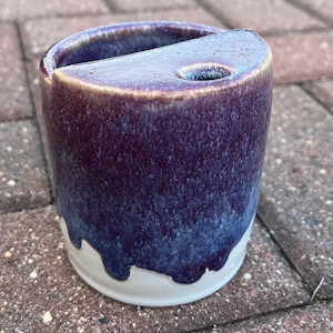 May include: A purple and white ceramic mug with a drip glaze. The mug has a small hole in the side for drinking.