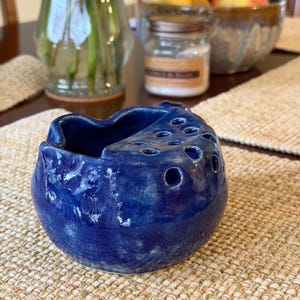 Puede incluir: Una taza de cerámica blanca con un asa negra y el texto "But First, Coffee" en negrita negra. La taza se encuentra sobre un fondo neutro, resaltando su diseño simple.