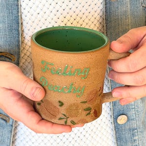 May include: A handmade ceramic mug with a speckled brown exterior and a green interior. The mug features the words "Feeling Peachy" in green script, along with painted leaf and peach designs. The mug has a curved handle.