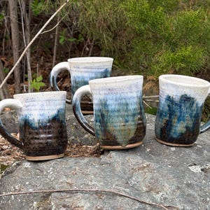May include: Four handmade ceramic mugs with a unique glaze. The mugs feature a gradient of colors, transitioning from dark brown at the base to shades of blue and white at the top. Each mug has a sturdy handle.
