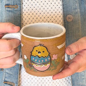 May include: A ceramic mug with a brown exterior, featuring a hand-painted design of a yellow chick hatching from a blue and pink egg. The mug has a white handle and rim, with floral accents. The mug is held by a person.